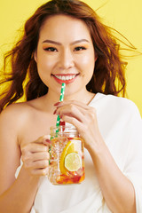 Fototapeta premium Portrait of Asian young woman with red hair holding glass and drinking cold lemonade from drinking straw on yellow background