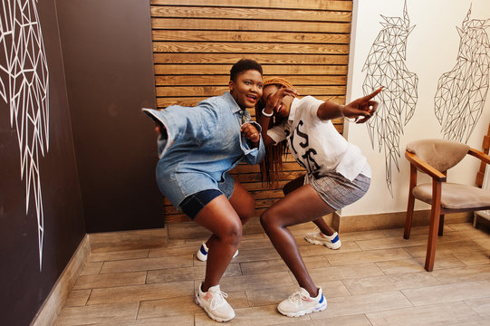 Two african womans in stylish casual clothes posing against wooden wall sat down and dance twerk.