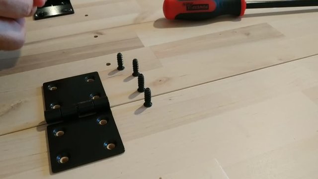 Fixing with a black metal hinge on two light wooden boards. One hand holds the hinge in place and the other positions the screws. 25Fps