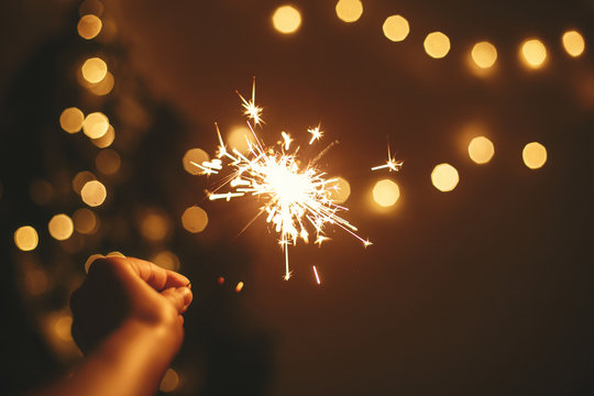 Happy New Year. Glowing Sparkler In Hand On Background Of Golden Christmas Tree Lights, Celebration In Dark Festive Room. Space For Text.   Fireworks Burning In Hand. Happy Holidays