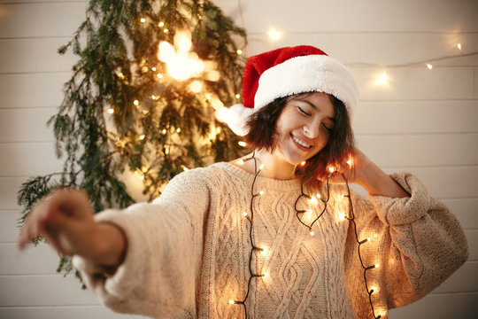  Stylish happy girl in santa hat holding sparkler on background of modern christmas tree lights in festive room. Happy New Year eve party. Hipster woman with fireworks. Happy Holidays