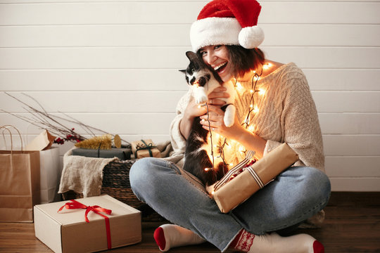 Stylish happy girl in santa hat playing with cute cat in festive christmas lights on background of modern room with presents. Young hipster woman in cozy sweater hugging kitty and smiling