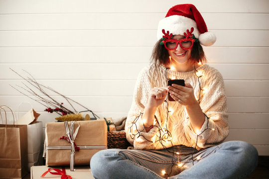 Stylish happy girl in festive glasses with reindeer horns holding phone and smiling in christmas lights. Young hipster woman in santa hat holding smartphone and laughing in christmas room