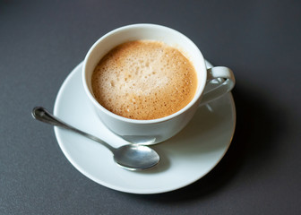 White cup of coffee with froth on dark gray background.