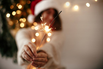 Happy New Year eve party atmosphere. Sparkler burning in hand of stylish girl in santa hat on...