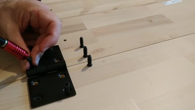 Fixing with a black metal hinge on two light wooden boards. A hand positions the screws. The screwdriver is used for screwing. 25Fps