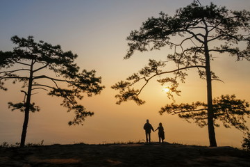 Mountain view Silhouette couple tourists standing on top hill under pine tree with yellow sun light in the sky background, sunrise at Nok Aen Cliff, Phu Kradueng, Loei, Thailand.