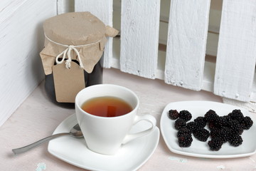 Jars of jam, covered with paper and tied with twine. On the string hang craft labels. Next to it is a plate of fresh blackberries and a cup of tea.