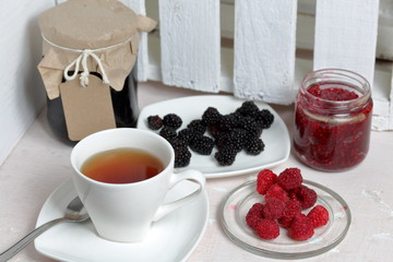 Jars of jam, covered with paper and tied with twine. On the string hang craft labels. One jar is open. Next to a plate of fresh raspberries, blackberries and a cup of tea.
