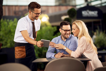 Happy waiter showing the menu on touchpad to his guests in a cafe.