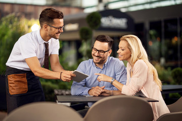 Happy couple and waiter looking at menu on digital tablet in a cafe.