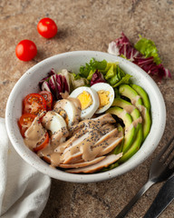 Top view of traditional american cobb salad with avocado, chicken breast, vinaigrette, tomatoes and hard boiled egg, served on a marble table with different ingredients around