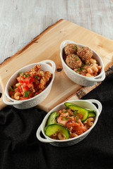 Different kinds of mac and cheese, served in portions on a wooden board: pasta with salmon and cucumber, pasta with meatballs and cheese, with chicken strips and tomatoes