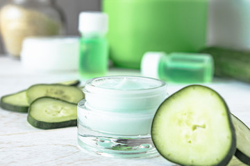 Face cream in a glass jar next to slices of fresh cucumbers on a white wooden background.