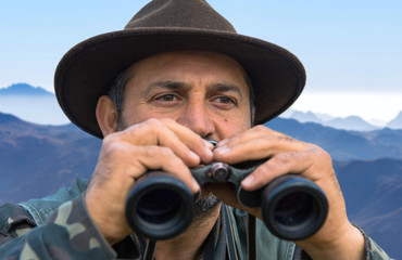 A hunter in a hat with binoculars looks out for prey.