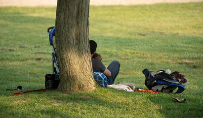 father takes a break under a shade tree at the park to hold his son and rest