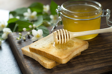wooden spoon with honey is lying on a piece of bread near a glass jar of honey