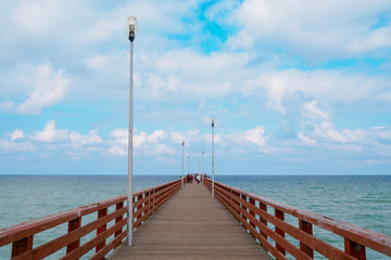 Sea piercing with a pier equipped with wooden railings and lanterns. Blue sky with white cumulus clouds.