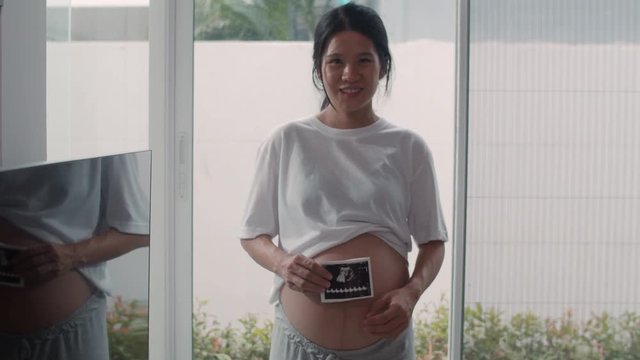 Young Asian Pregnant women show and looking ultrasound photo baby in belly. Mom feeling happy smiling peaceful while take care child lying near window in living room at home concept. Slow motion.