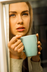 Thoughtful young blond woman with cup of coffee looking through the window.