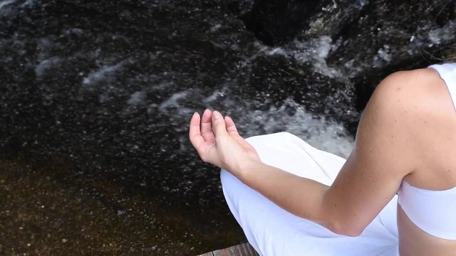 Young woman is practicing yoga and meditation in lotus pose at waterfall, camera moves from left to right in slow motion