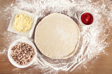 Raw pizza dough on a very floured wooden table. Around it are the ingredients that will be added to the pizza