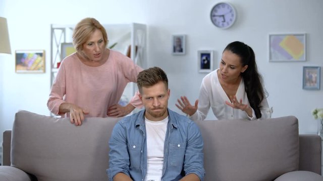 Tired man sitting on sofa, enduring to wife and mother criticize him, quarrel