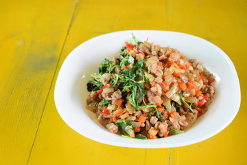 Spicy Thai food, stir-fried pork basil in a white dish placed on a yellow wooden table.