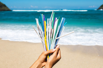 Hand holding heap of used plastic straws on background of clean beach and ocean waves. Plastic ocean pollution, environmental crisis. Say no plastic. Single-use plastic waste