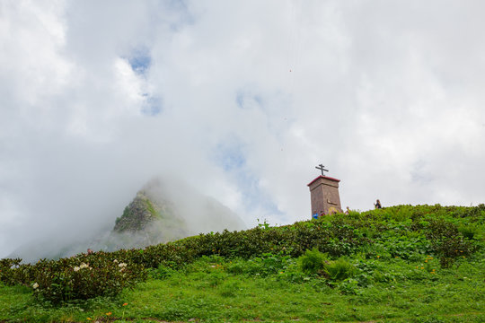 A small Christian chapel on a hill with bright green plants, a mountain behind, a blue sky in white clouds, people on a hill, white flovers in down left corner. Day light, horizontal image.