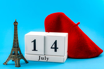 Happy Bastille day and French national day concept with a red beret, a block calendar set on July 14, a miniature of the Eiffel Tower isolated on blue background