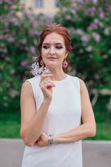 Charming woman wearing beautiful white dress standing next to lilac bush