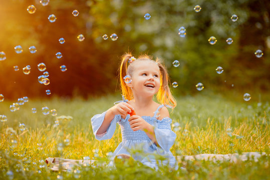 A little girl blows soap bubbles on a green lawn in the rays of a sunset