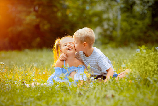 Beautiful children sit on a green lawn in the rays of a sunset. The boy hugs and kisses the girl on the cheek. Happy children
