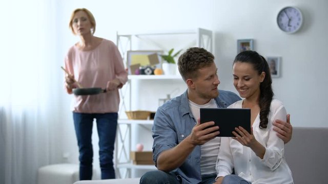Curious mother-in-law looking young couple scrolling on tablet PC at home