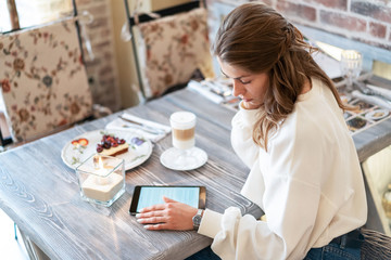 beautiful girl during breakfast in cafe reads e-book. Work on tablet during break. Freelance. Creative person. Latte coffee, cheesecake.