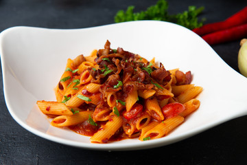 Penne pasta in tomato sauce with bacon on dark background