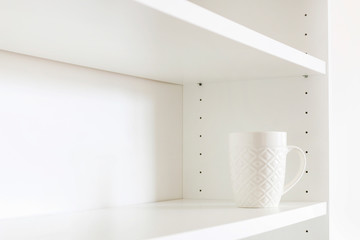 A white ceramic mug standing on an empty shelf.