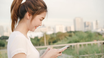 Cheerful Asian tourist blogger woman using touchscreen technology at smartphone while walking on the street at downtown city in the evening. Lifestyle backpack tourist travel holiday concept.