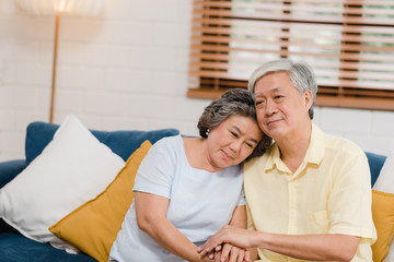 Asian elderly couple holding their hands while taking together in living room, couple feeling happy share and support each other lying on sofa at home. Lifestyle Senior family at home concept.