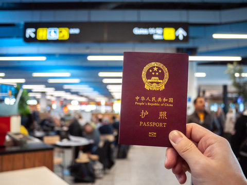 Hand holding a Chinese passport with airport foodcourt and direction sign as background. 