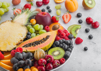 Fototapeta na wymiar Fresh raw organic summer berries and exotic fruits in white plate on light background. Pineapple, papaya, grapes, nectarine, orange, apricot, kiwi, pear, lychees, cherry and physalis. Macro