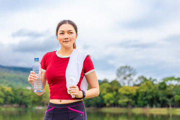 Beautiful Asian athlete woman drinking water during her break from morning  exercise at a lake park with view of lake and mountains in the background, healthy lifestyle