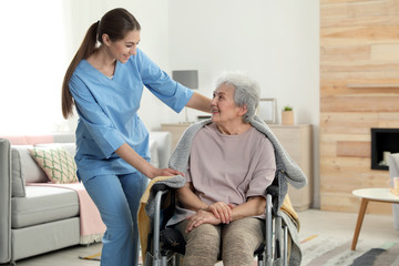 Nurse covering elderly woman in wheelchair with blanket indoors. Assisting senior people