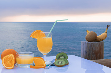 A ripe squeezed orange, green kiwi, orange juice, a freshly harvested whole kiwi and banana on a wooden dock - The sea under a cloudy sky