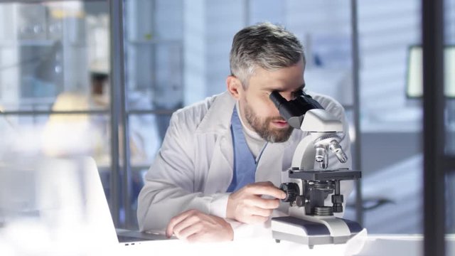 Panning waist-up shot of 40-something grey-haired Caucasian professor, wearing scrubs and lab coat, working on scientific research paper, analyzing tissue sample under microscope and typing on laptop