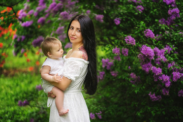 Young mother holds her little daughter in her arms among blooming trees. Lilac tree at background