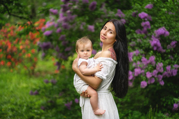 Cute Baby with big blue eyes with young beautiful mother at spring. Blooming lilac tree at the background.
