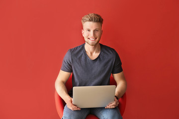 Handsome young man with laptop sitting on chair against color background