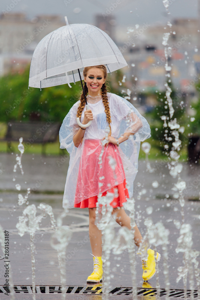 Wall mural young pretty girl with two braids in yellow boots and with transparent umbrella stands near fountain